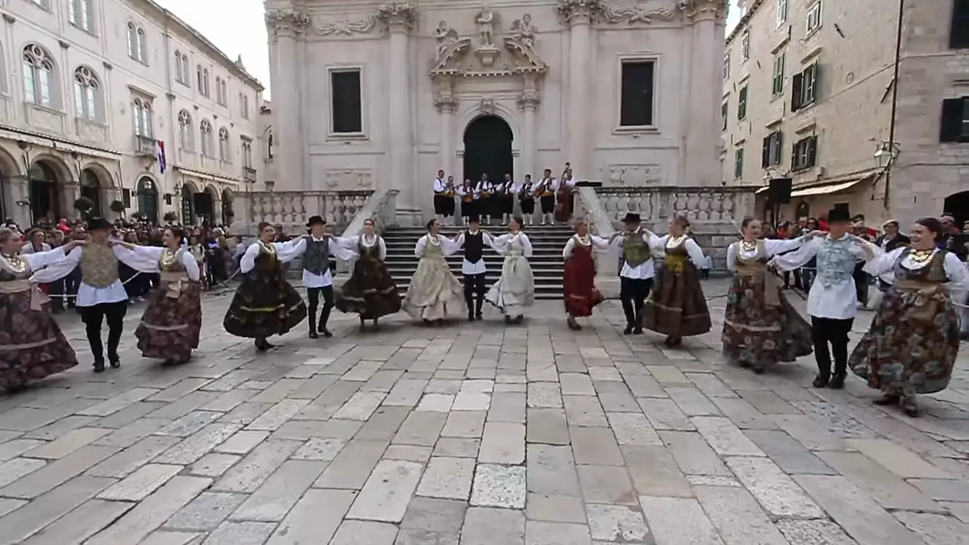Dansere i mørke broderede nederdele, hvide bluser og veste danner en linje over en stensat plads i Dubrovnik, med et orkester på trappen foran en barokkirke bag dem.