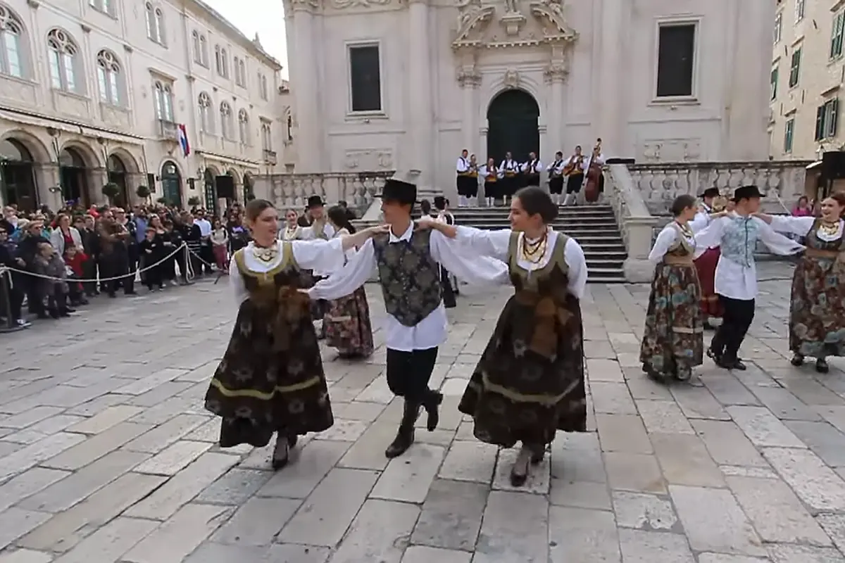 To kvinder i mørke broderede nederdele og en mand i vest og sort hat danser hånd i hånd på brolægningen med musikanter på trappen foran en barokkirke bag dem.